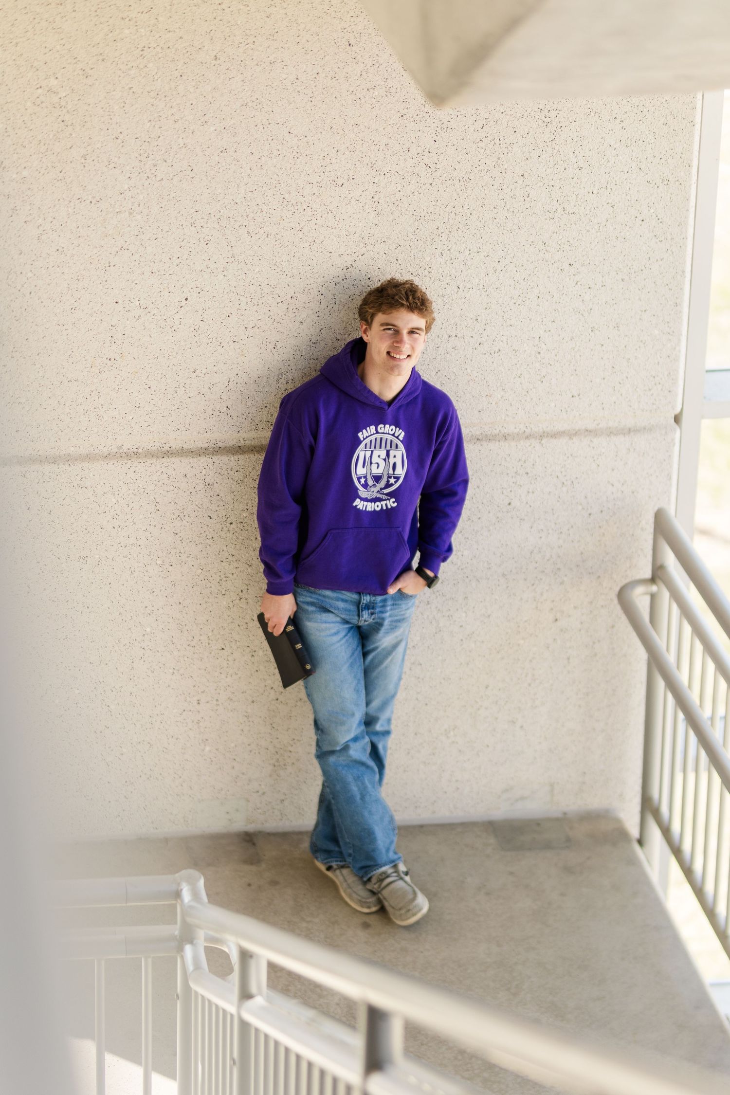 Person wearing purple collegiate sweatshirt and jeans standing against white institutional wall.