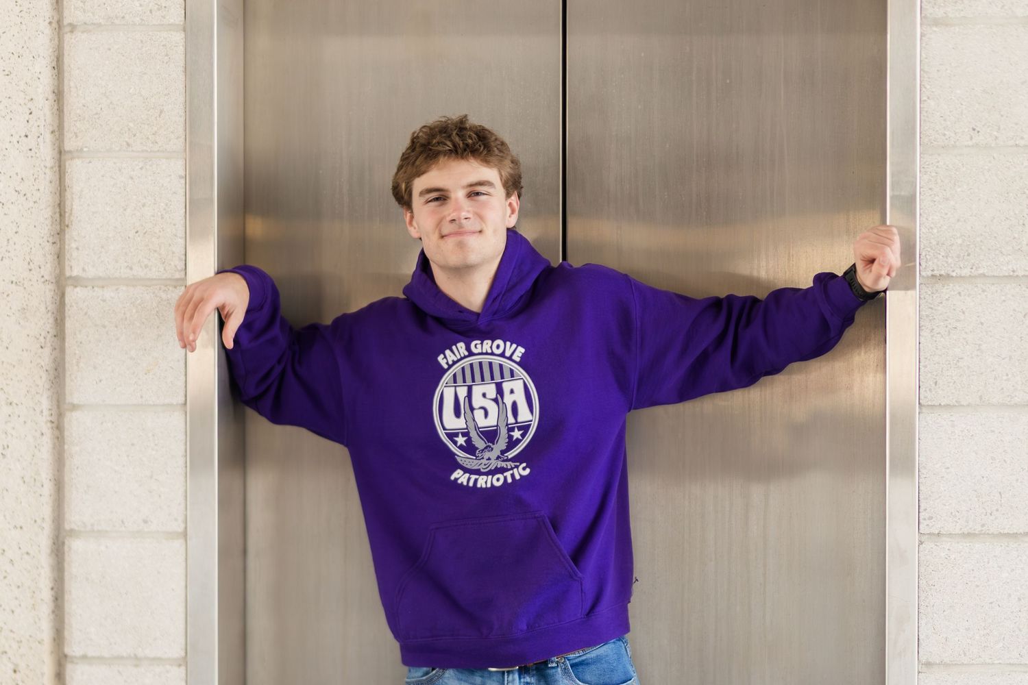 Young person wearing purple USA sweatshirt poses against elevator door and wall in casual stance.