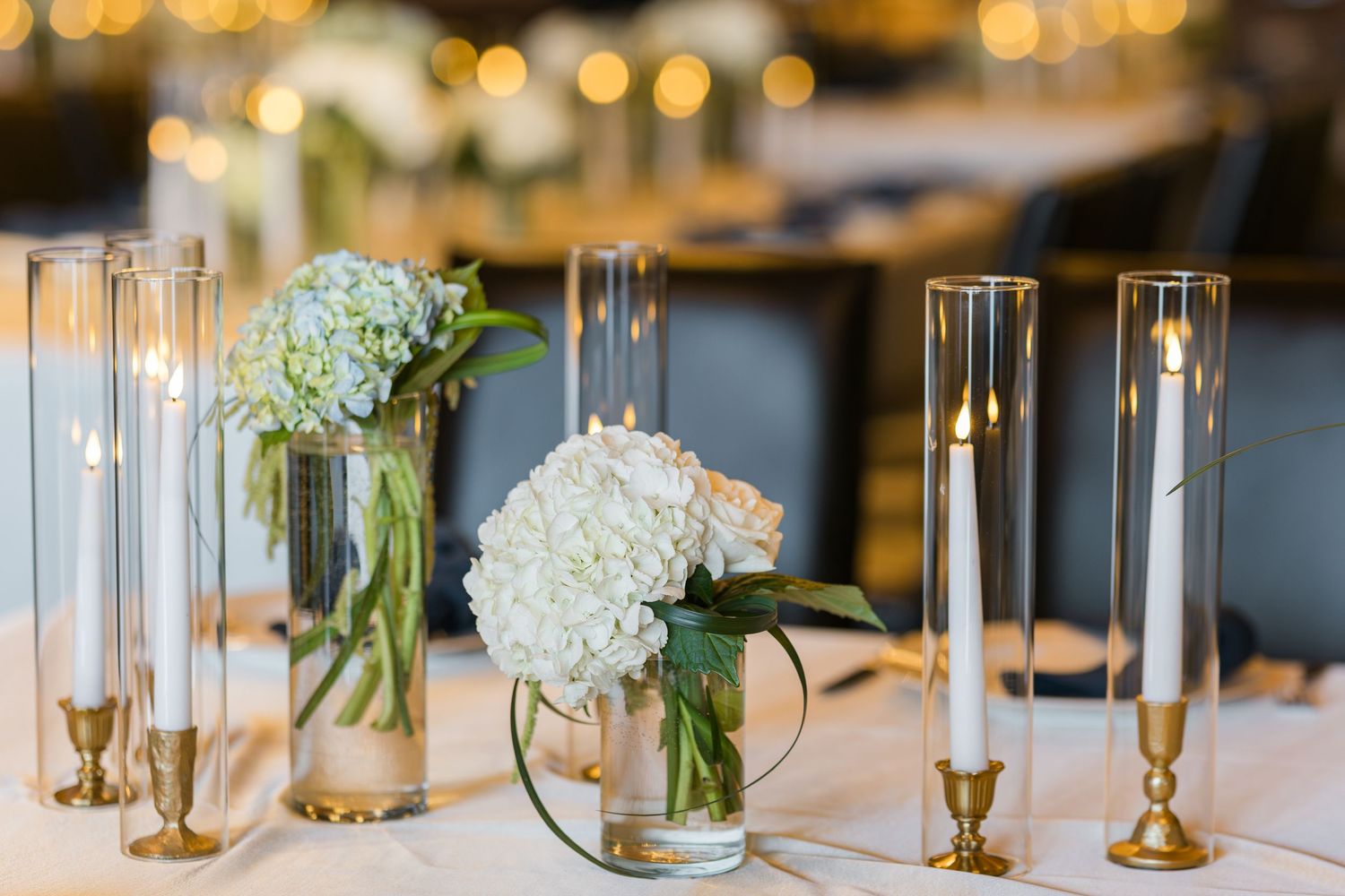 Elegant wedding reception tables decorated with white hydrangeas, tall glass candleholders and golden accents.