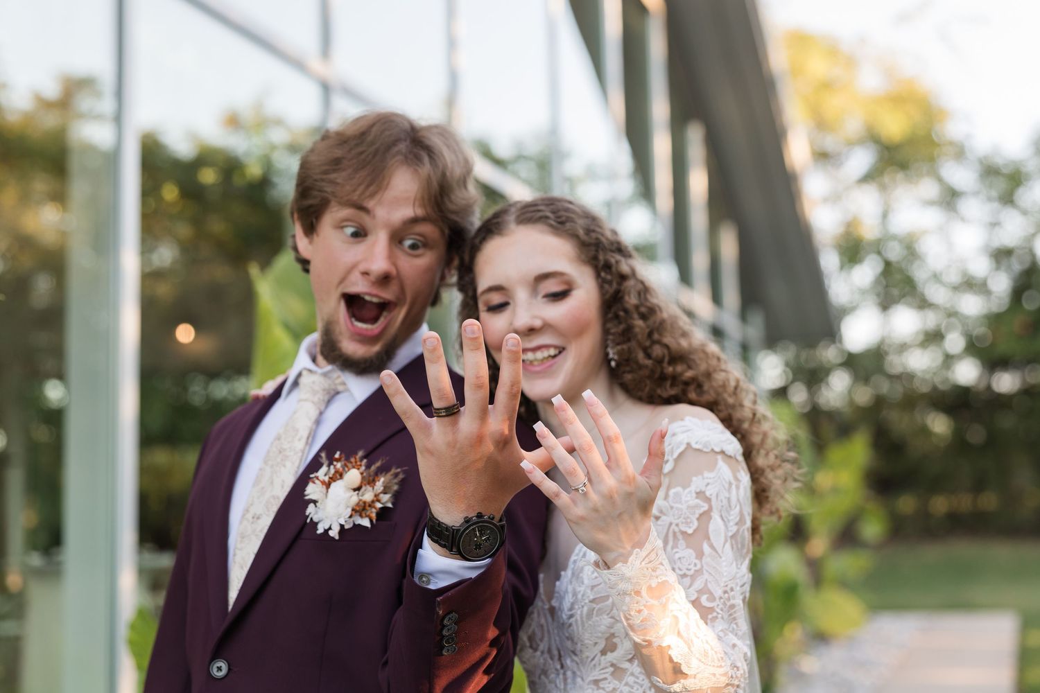 Joyful newlyweds showing off wedding rings, groom in burgundy suit with boutonniere, bride in lace dress, outdoors.