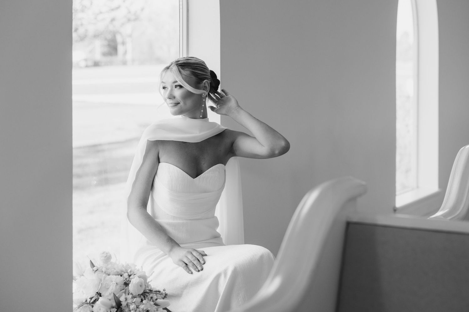 Black and white photo of an elegant bride in a strapless gown sitting by a window, gazing outside on her wedding day.