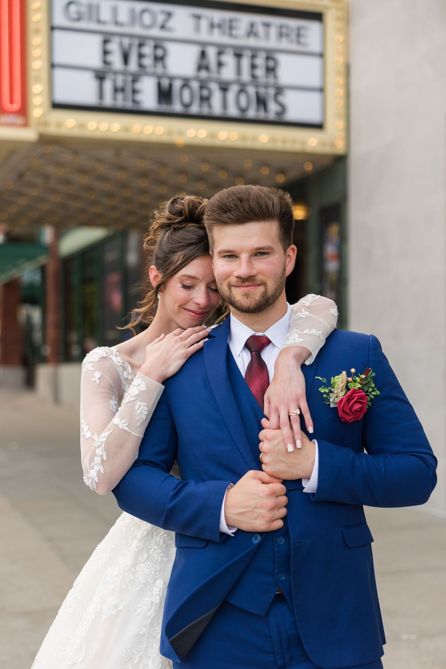 Bride in lace gown embraces groom in navy suit outside Gillioz Theatre Ever After The Mortons sign.