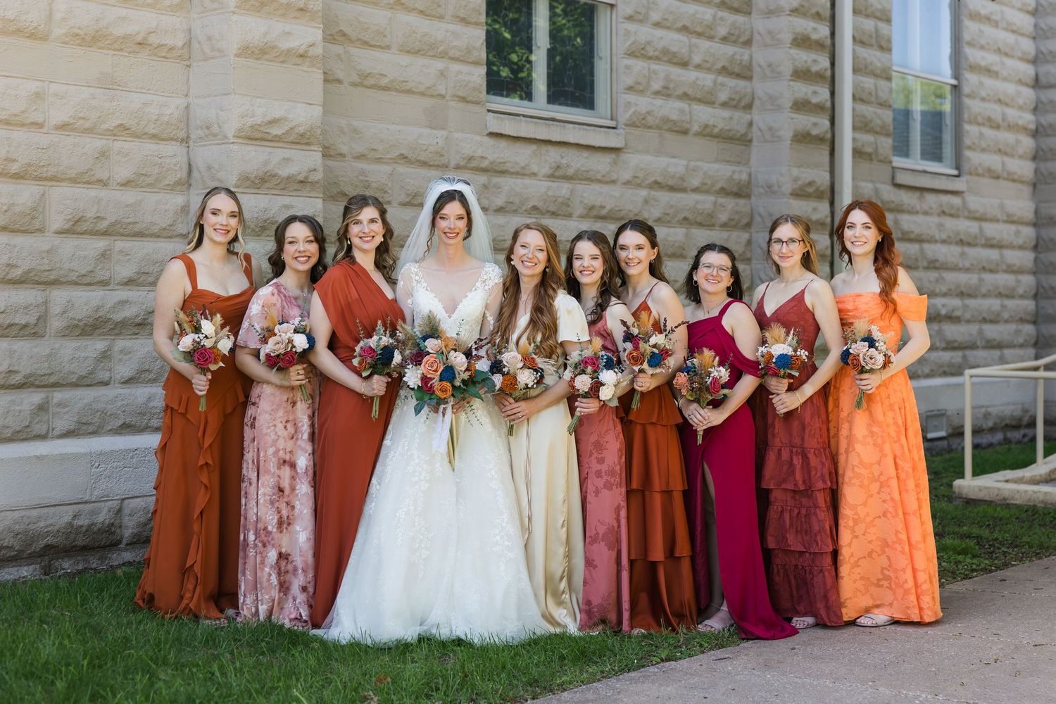 Bride with bridesmaids in rust and orange dresses holding colorful bouquets outside a stone building.