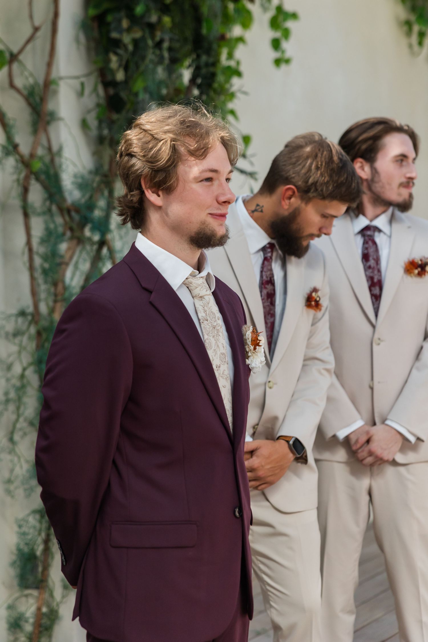 Groom in burgundy suit standing with groomsmen in beige suits at wedding ceremony altar.