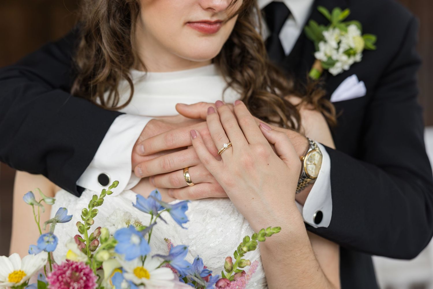 Bride and groom holding hands showing wedding rings, bride holding colorful bouquet with blue and pink flowers.