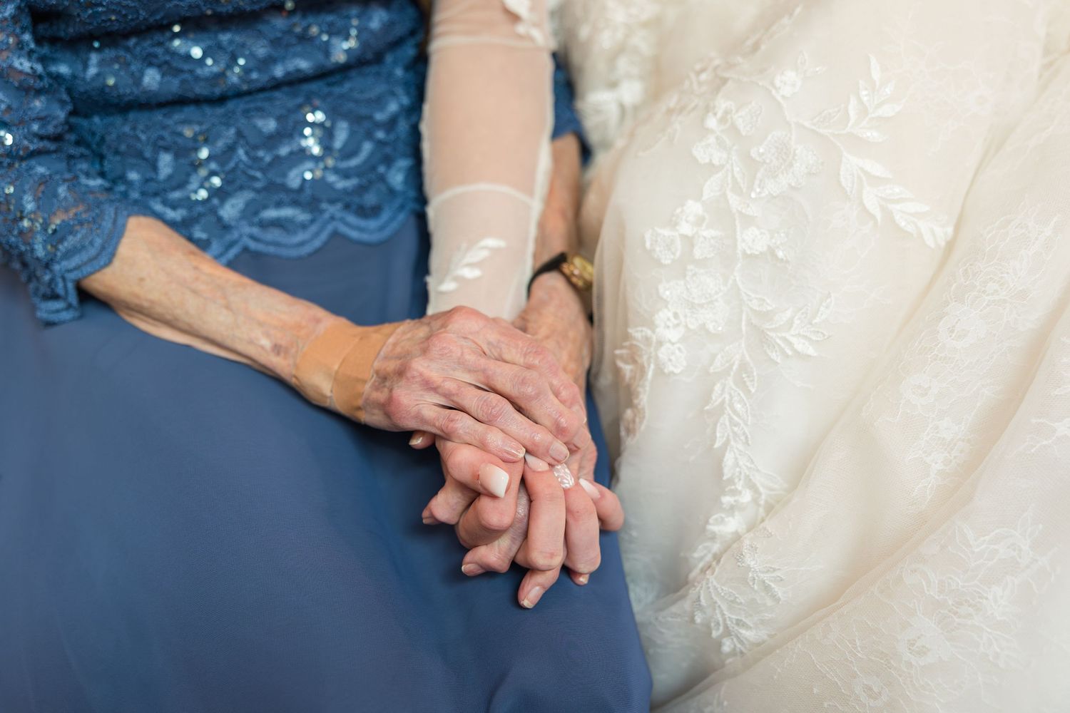 Elderly woman and bride holding hands together, wearing blue lace dress and white wedding gown at a wedding ceremony.