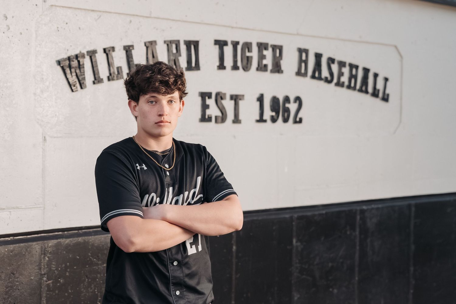 Baseball player poses against Willard Tiger Baseball wall in black uniform.