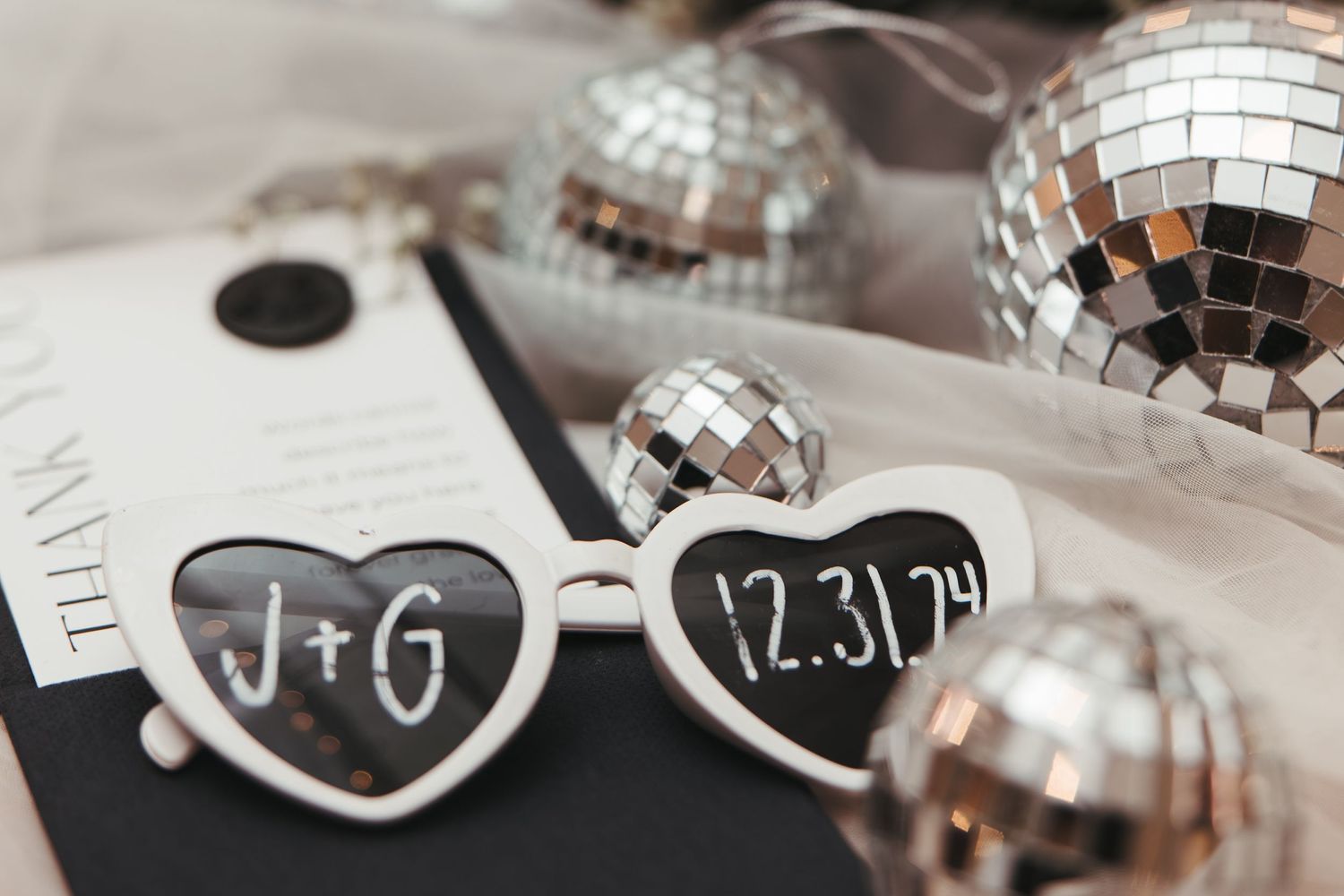 Heart-shaped sunglasses with date and disco balls arranged with wedding invitation.
