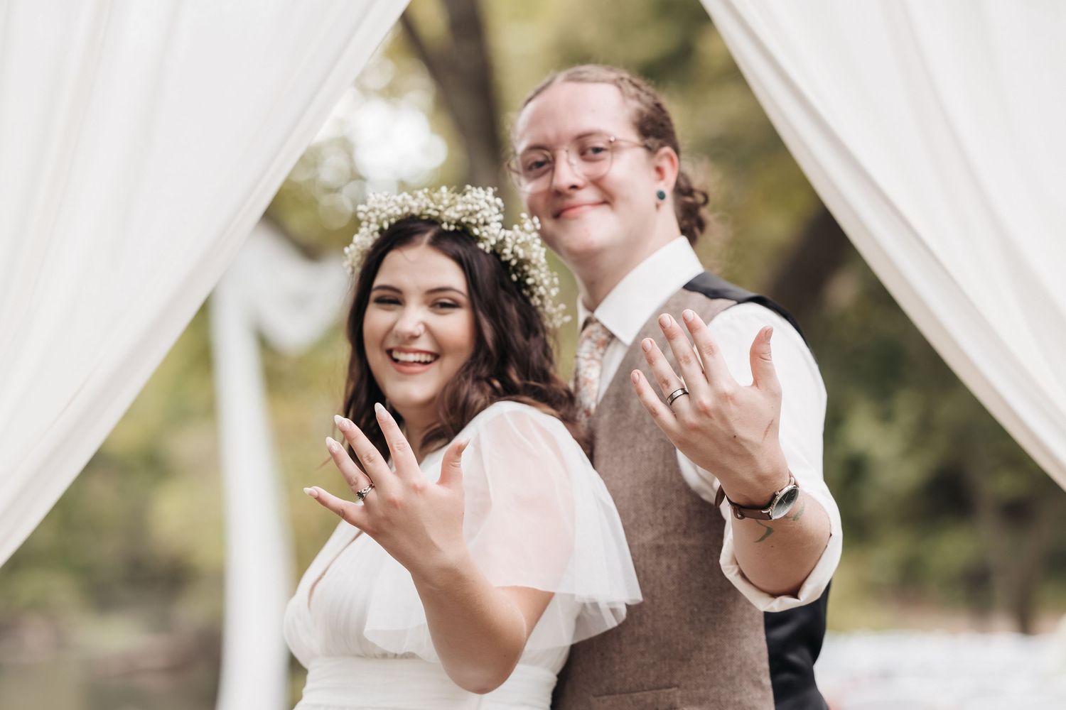 Wedding couple shows off their rings under white fabric arch outdoors.