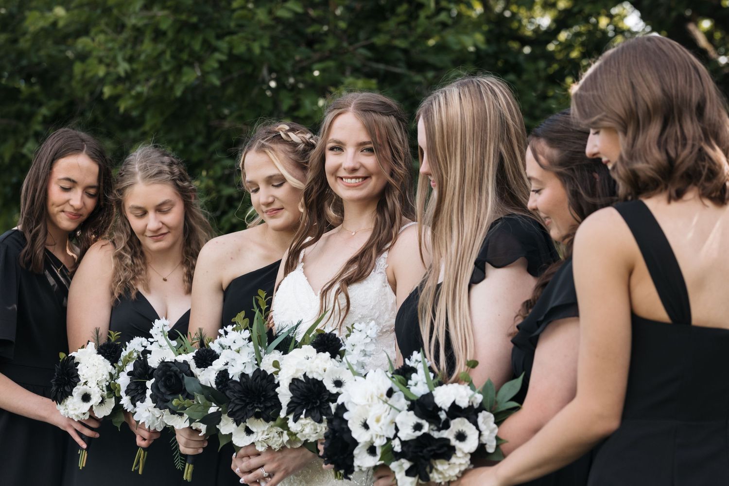 Bridesmaids in elegant black dresses hold white bouquets while standing together outdoors.