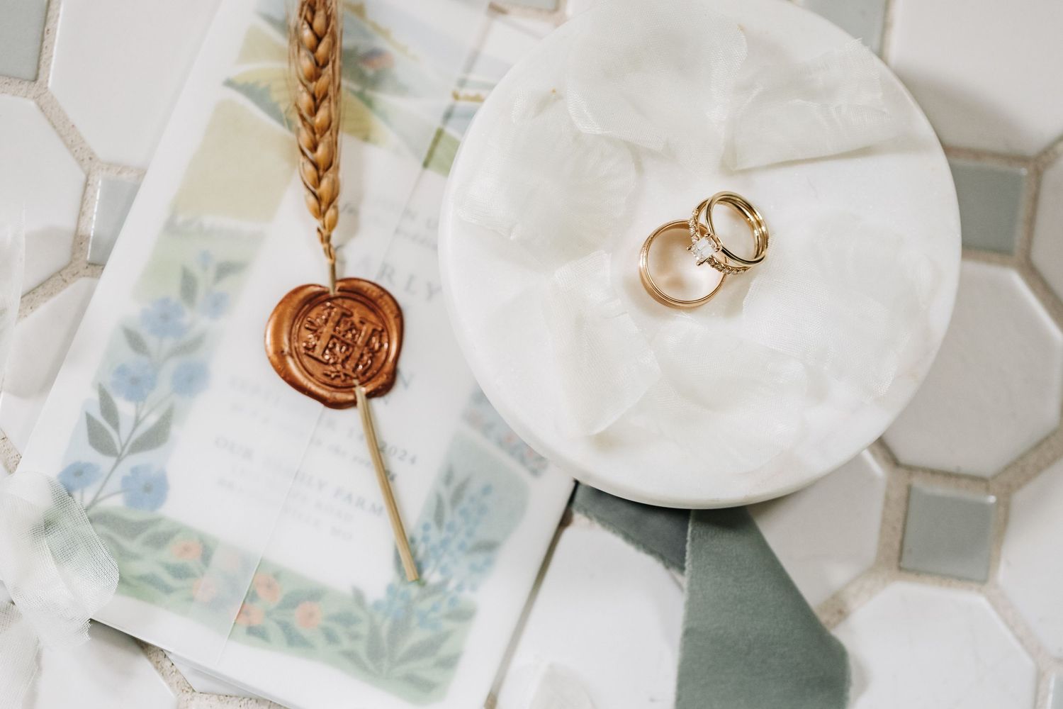 Wedding rings and a wax seal rest on white hexagonal tile with delicate blue floral accents.