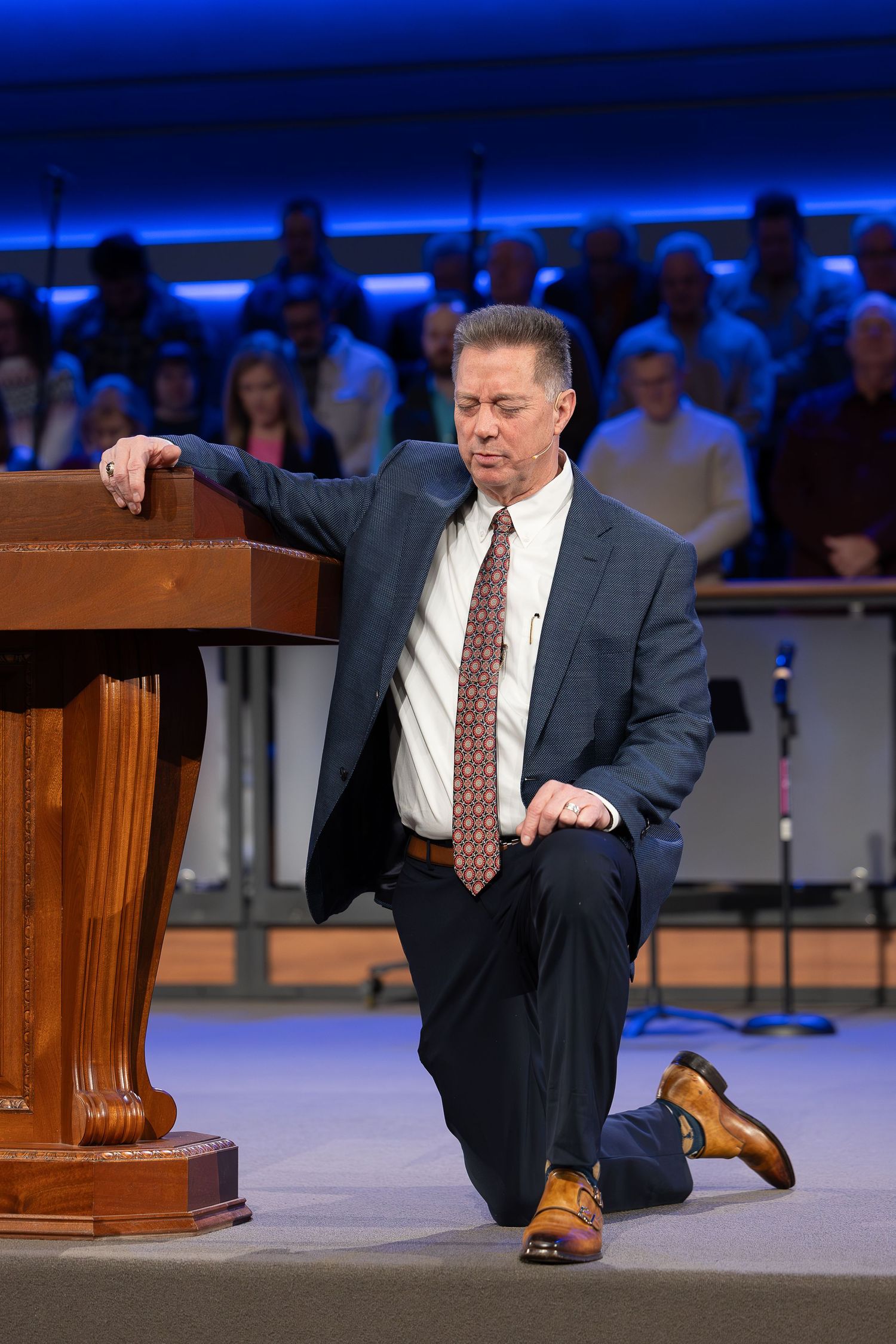 Speaker kneels at wooden podium on stage during presentation.