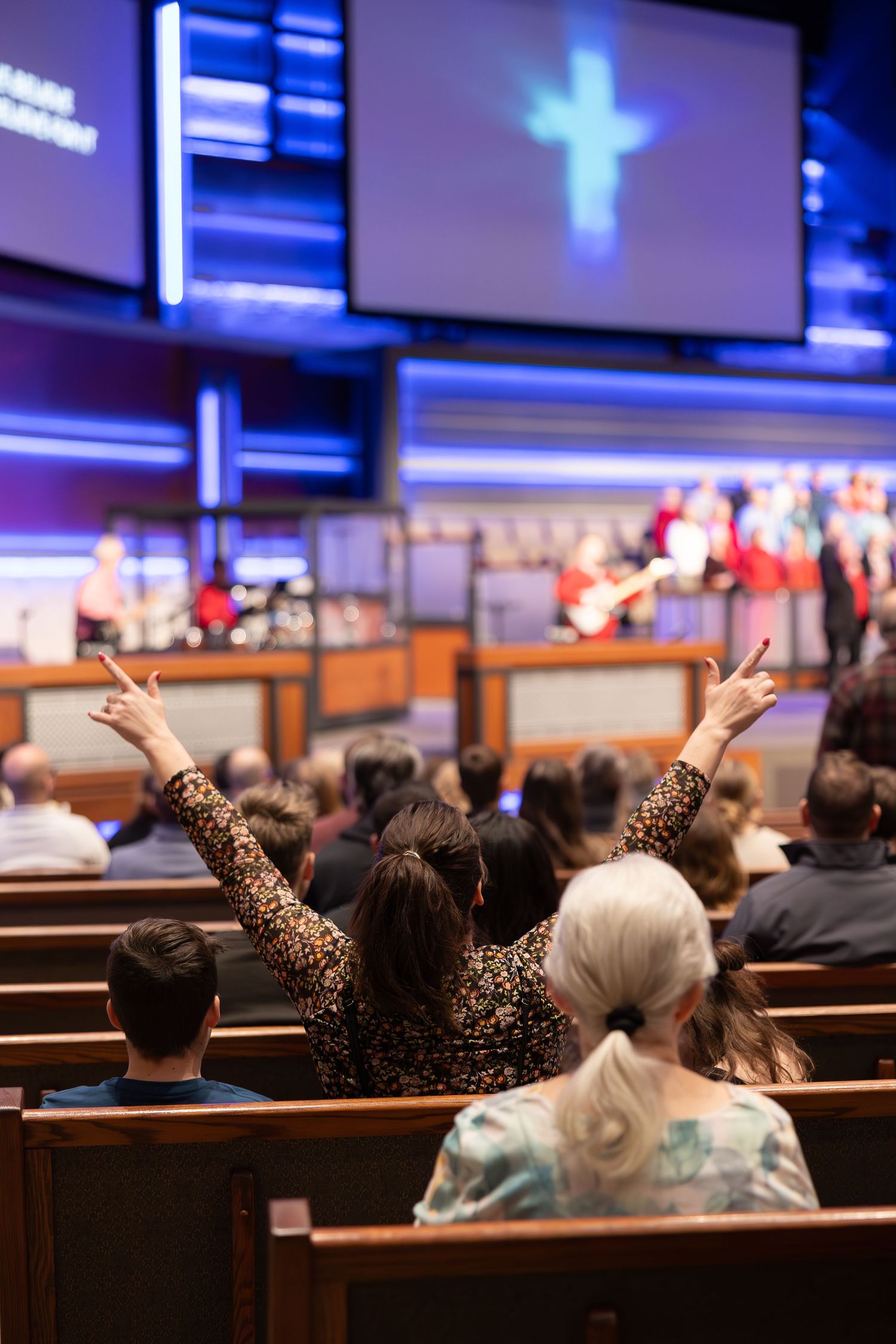 Congregation members raising hands during worship service with glowing cross on display screen.