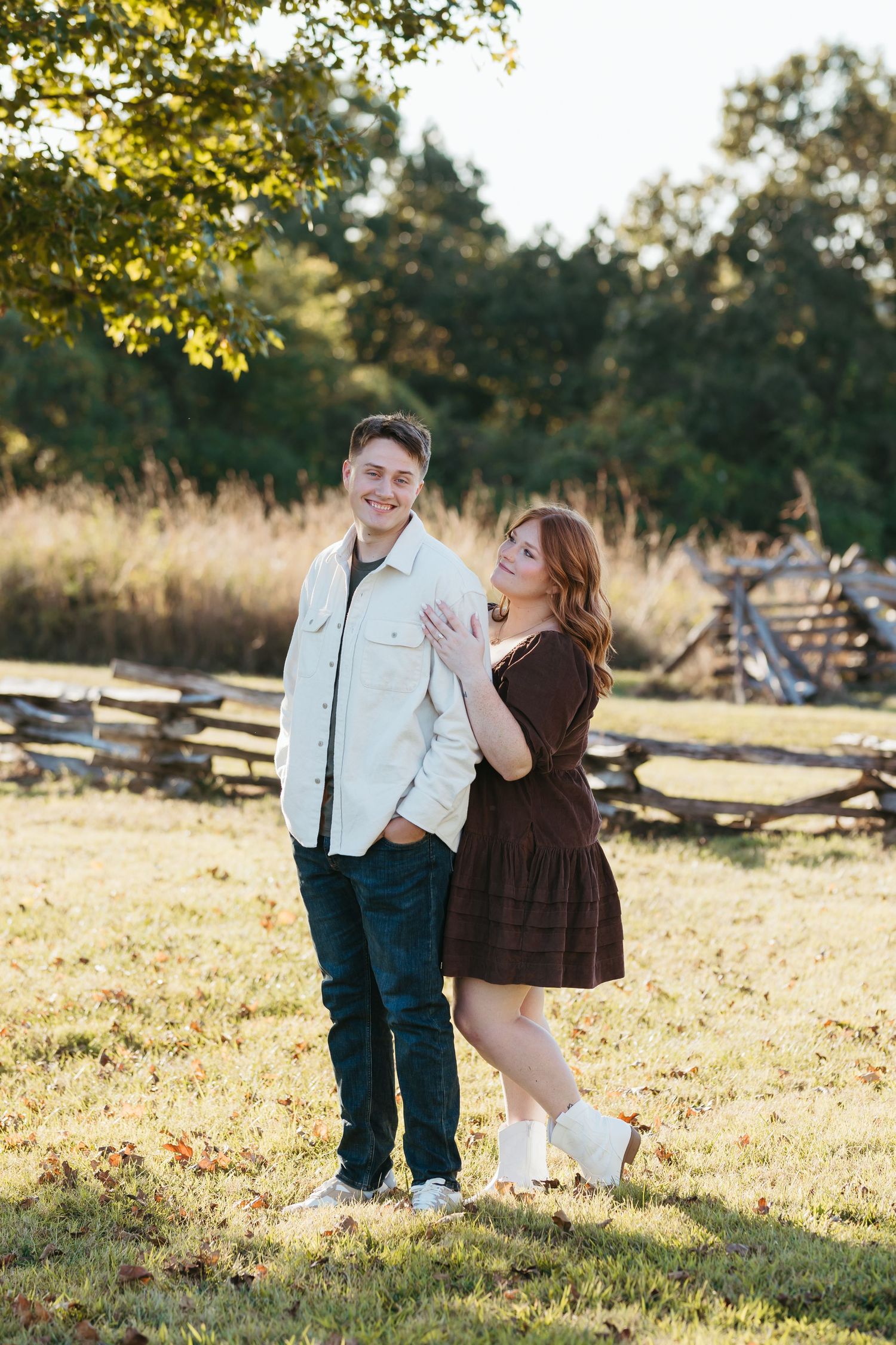 Couple laughing together in sunlit field with rustic fence in background.