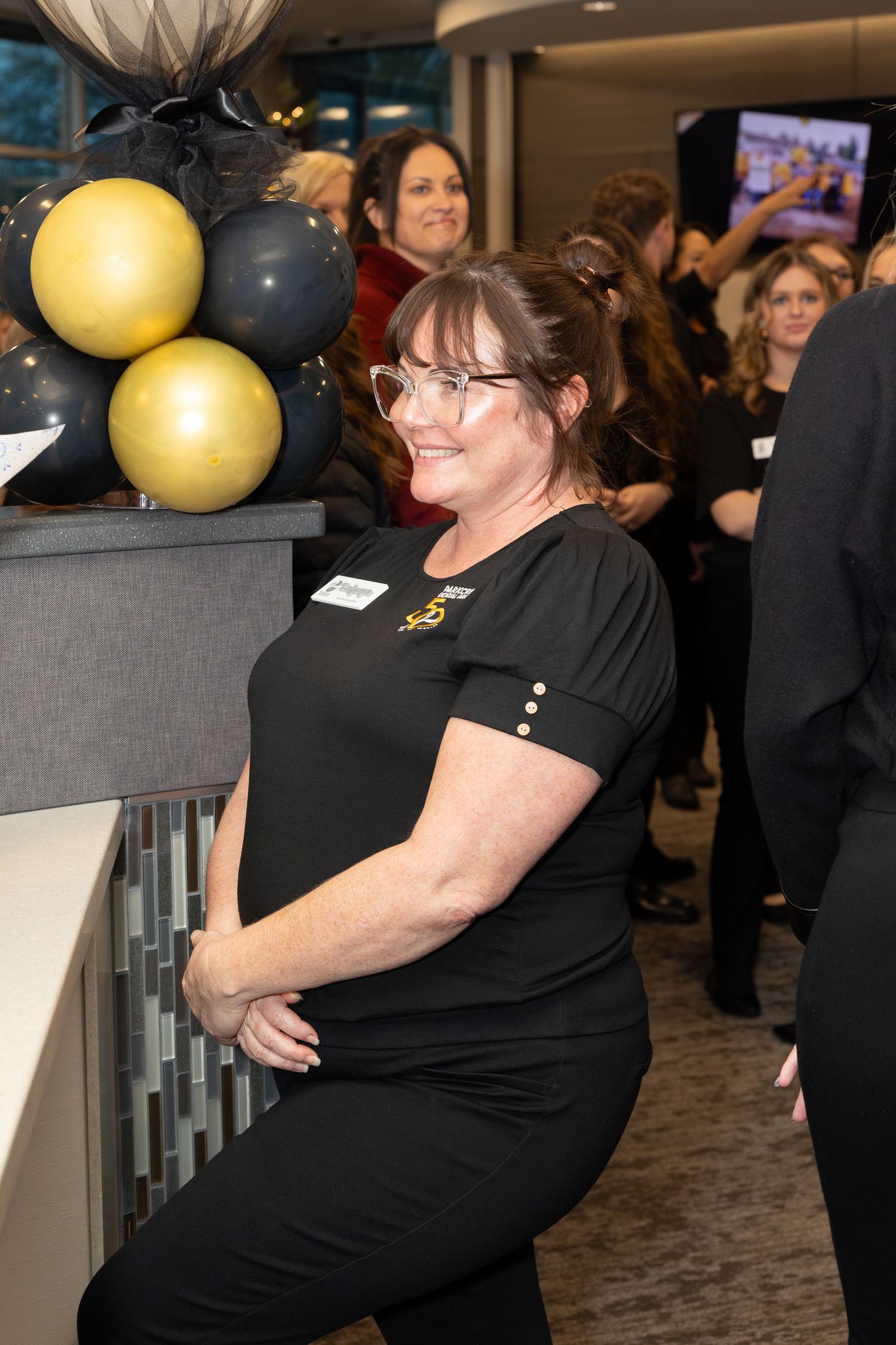 Staff member in black uniform poses during office celebration with gold and black balloons.