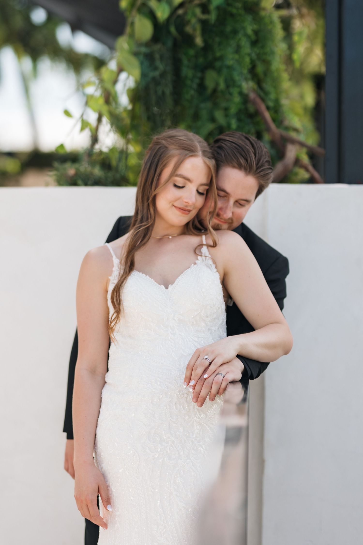 Romantic wedding portrait showing couple in intimate pose against white wall with hanging greenery.