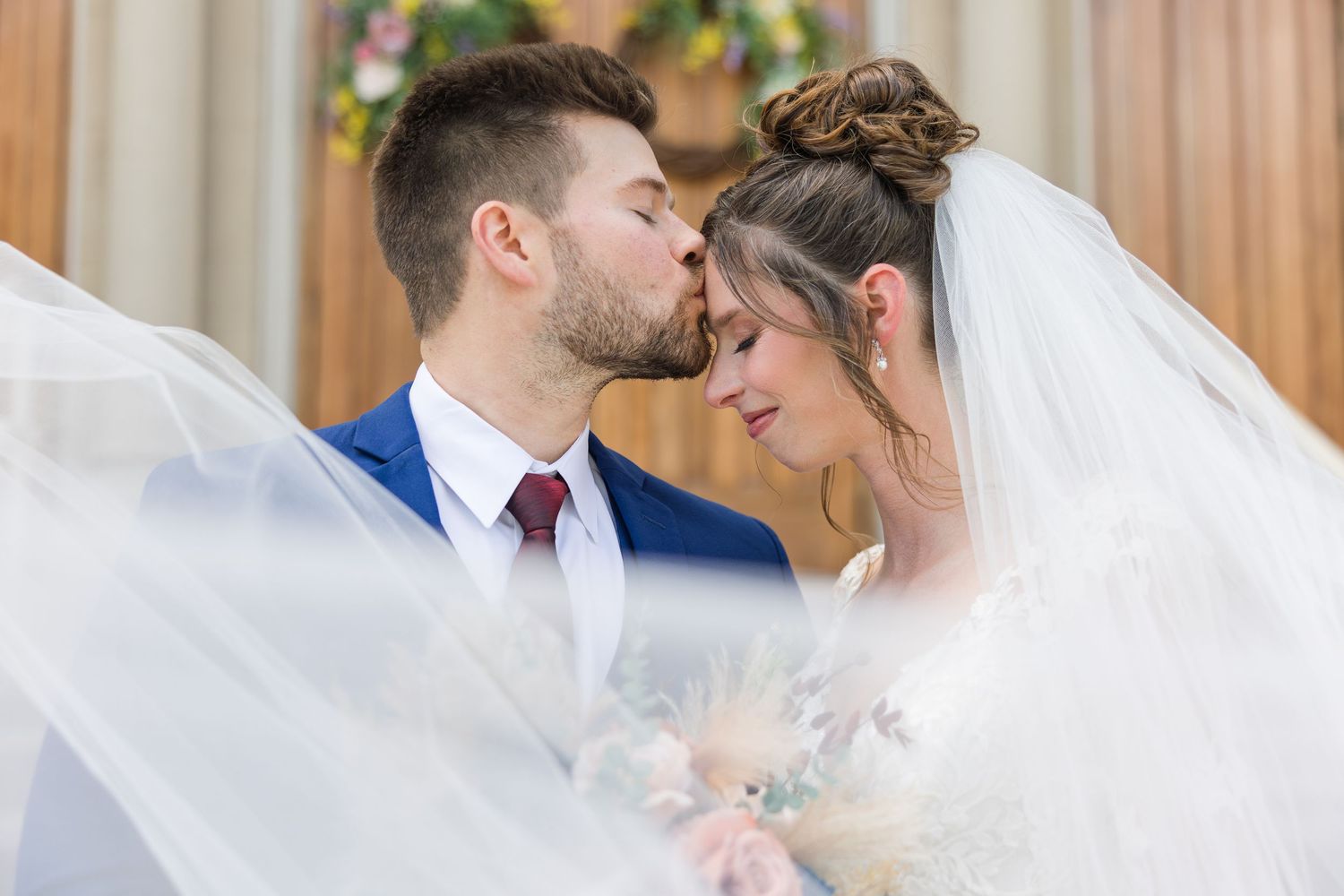 Groom kisses bride on forehead as flowing veil surrounds them during romantic wedding ceremony moment.