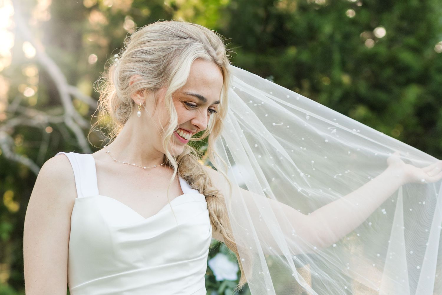Smiling blonde bride in white gown holding a pearl-dotted veil outdoors with soft green bokeh background.