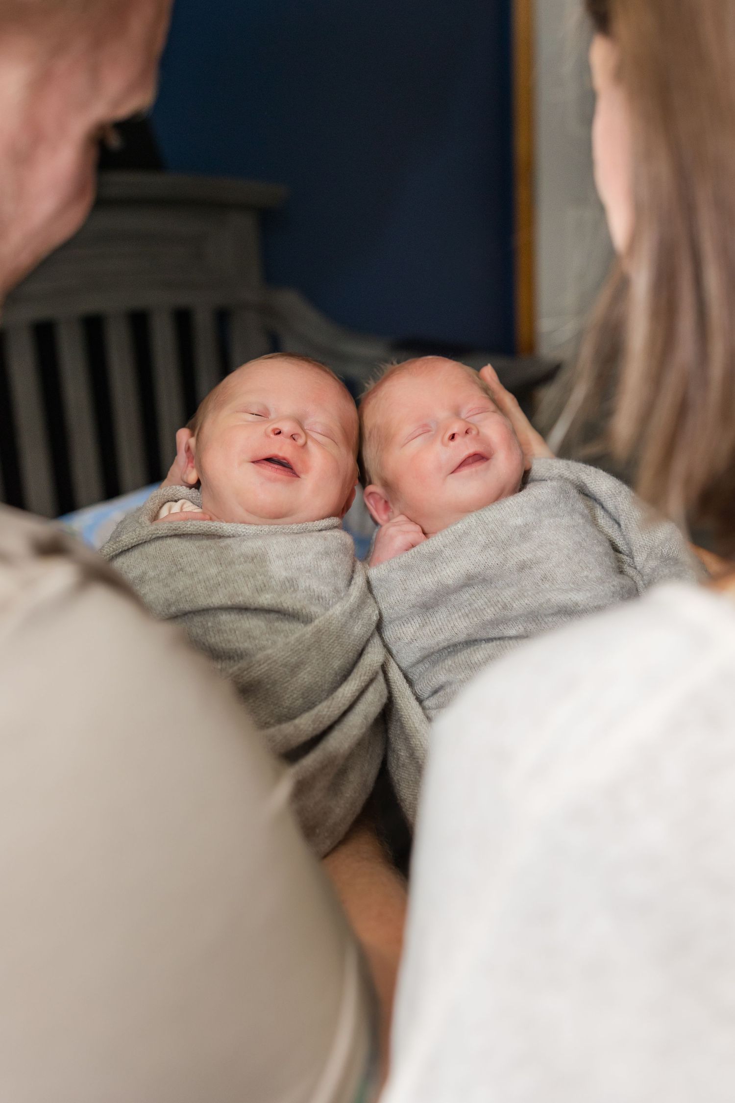 Two swaddled newborn twins smiling while being held by two adults near a crib.