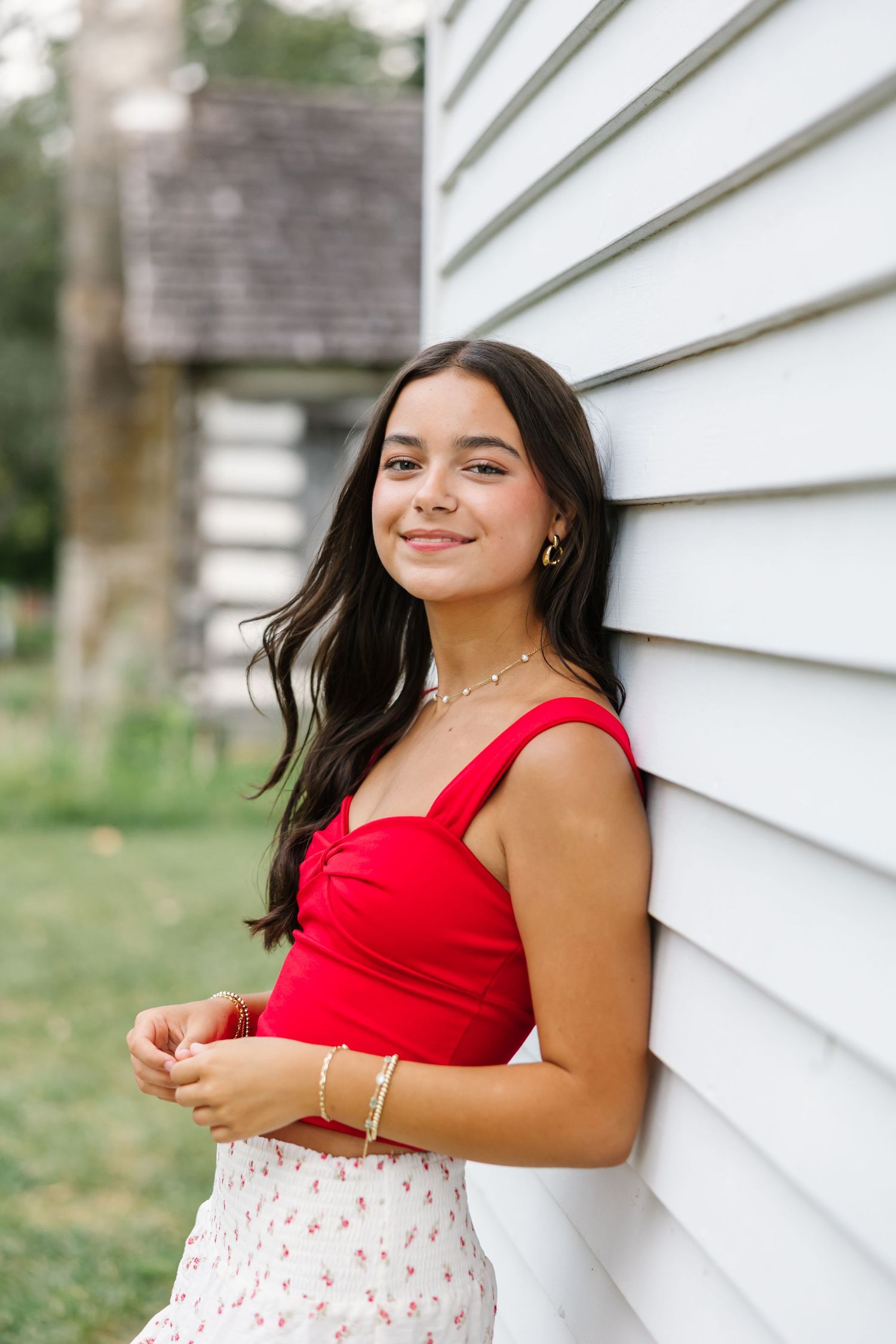 Young person in a red top and floral skirt poses against white siding in a candid outdoor photo shoot.