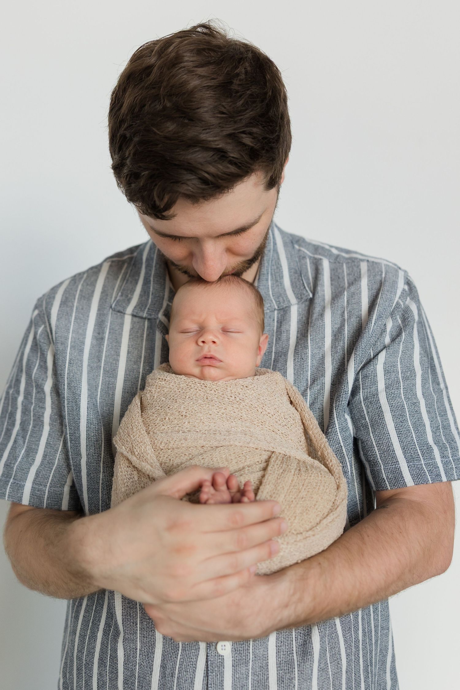 A tender moment captured as someone in a striped shirt gently holds and kisses a sleeping newborn wrapped in beige.
