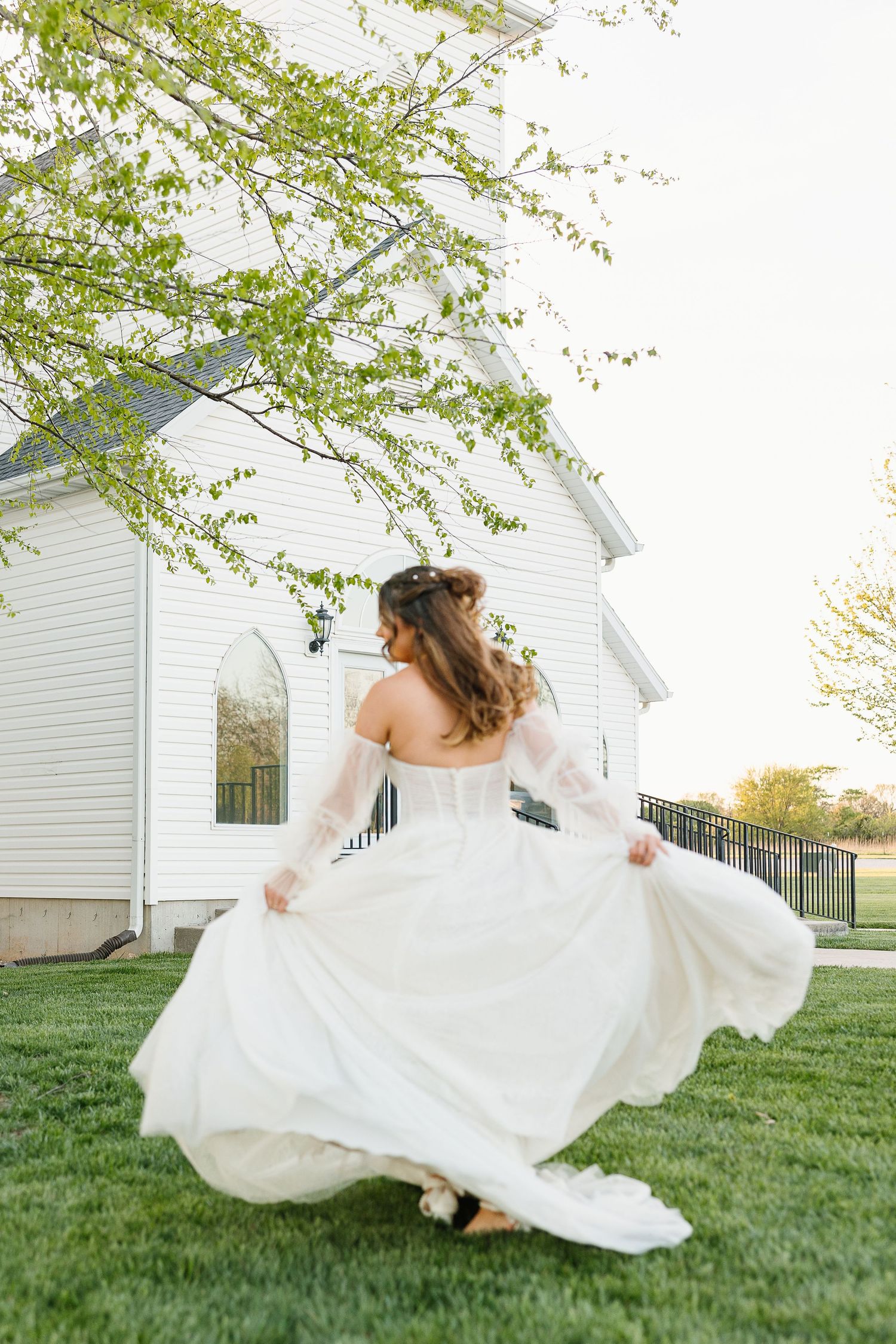 A flowing white wedding dress twirls elegantly on a green lawn in front of a white church building during springtime.
