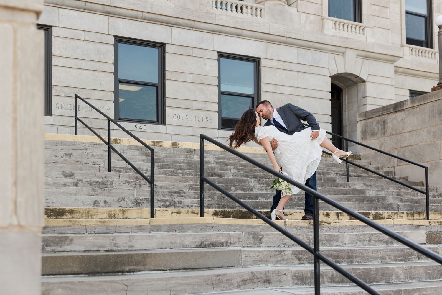 A couple shares a romantic dip pose on stone steps with black railings outside a classic stone building.