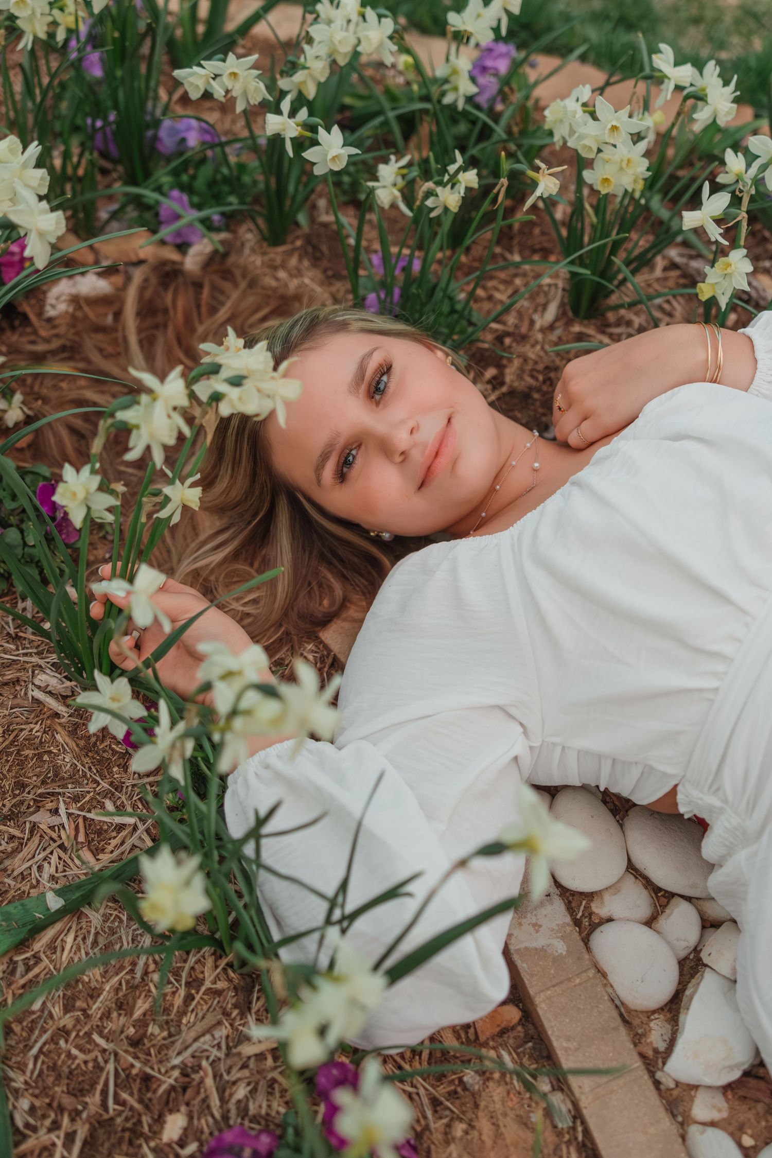 White daffodils and spring flowers bloom around a figure in a white dress resting on garden mulch and stones.