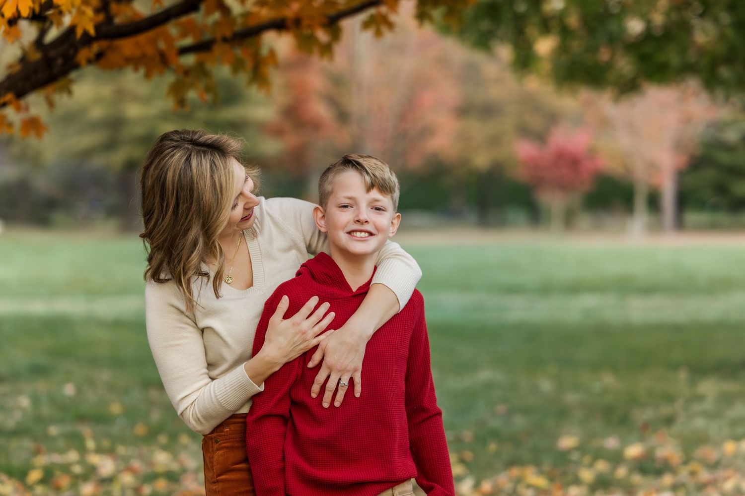 A couple in red and beige outfits embrace on grass during autumn with golden leaves in the background.