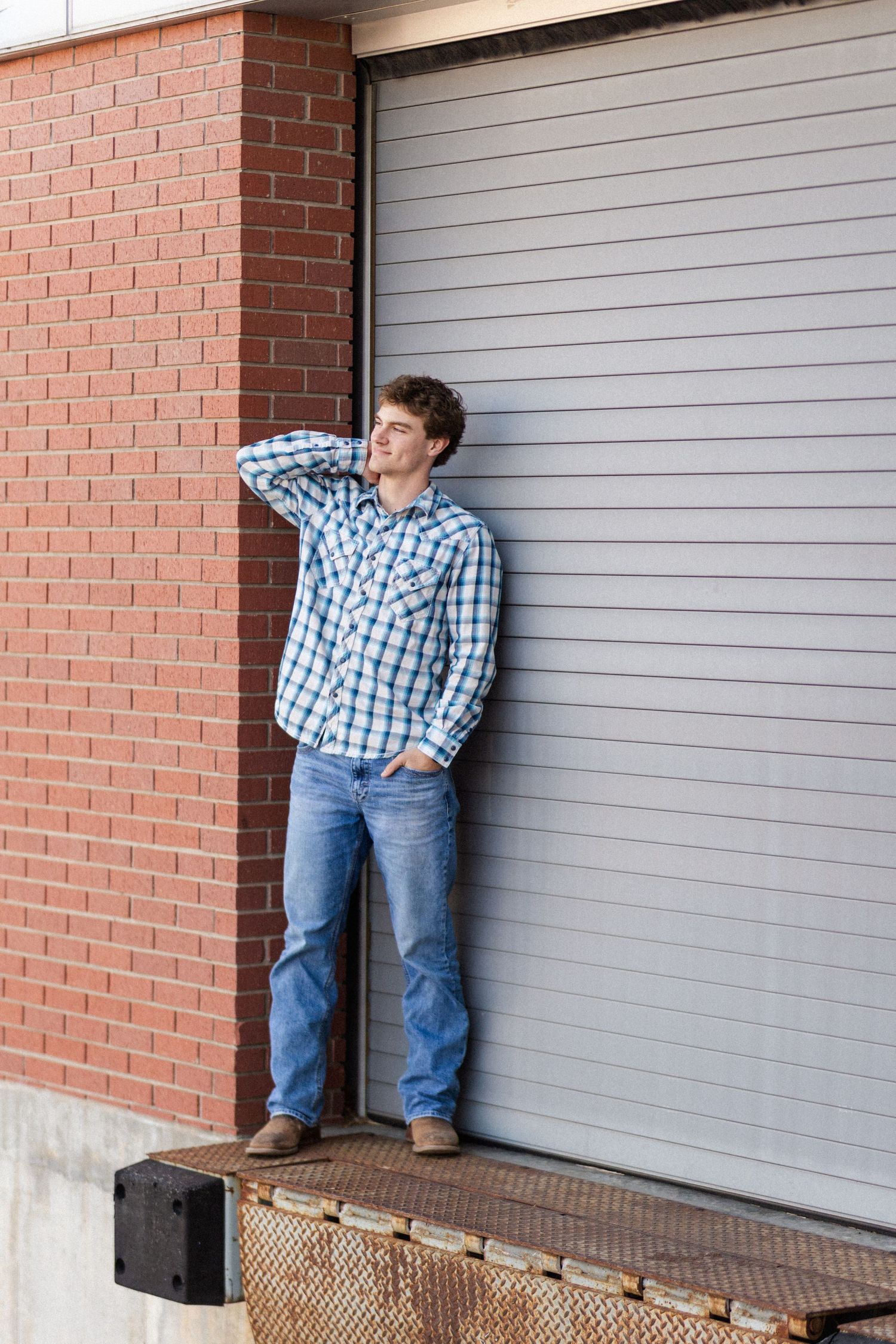 Person in plaid shirt and jeans poses casually against brick wall and loading dock door on warehouse dock.