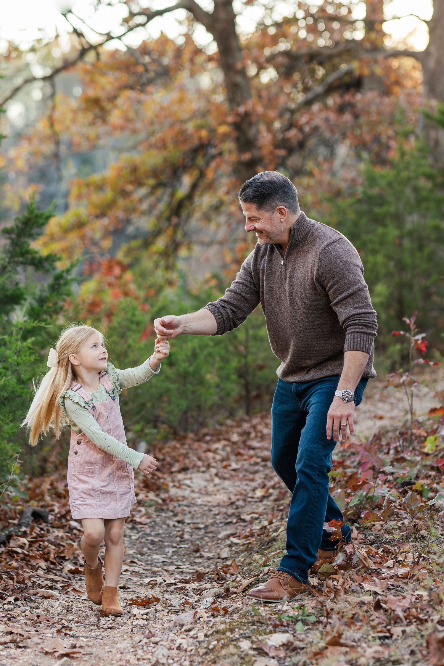 Adult and child holding hands while walking on autumn leaf-covered trail.