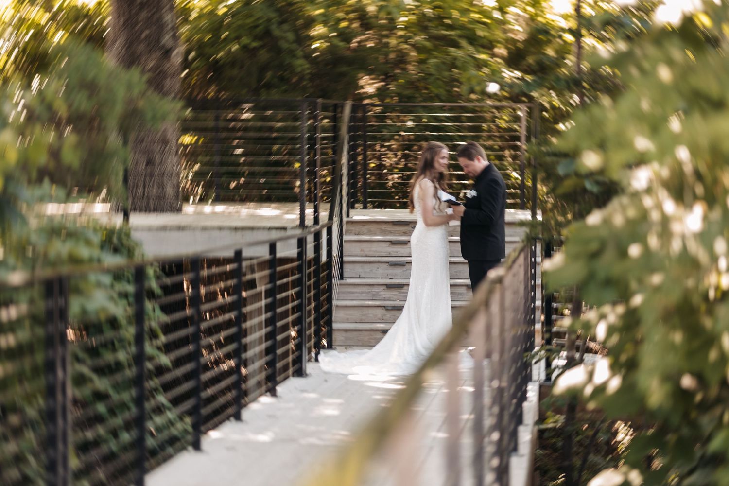 Wedding couple photographed from afar on elevated wooden walkway among trees.