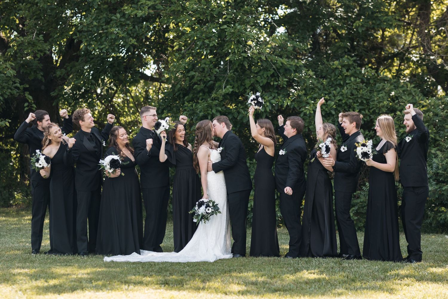 Large wedding party in formal black attire poses with bride outdoors.