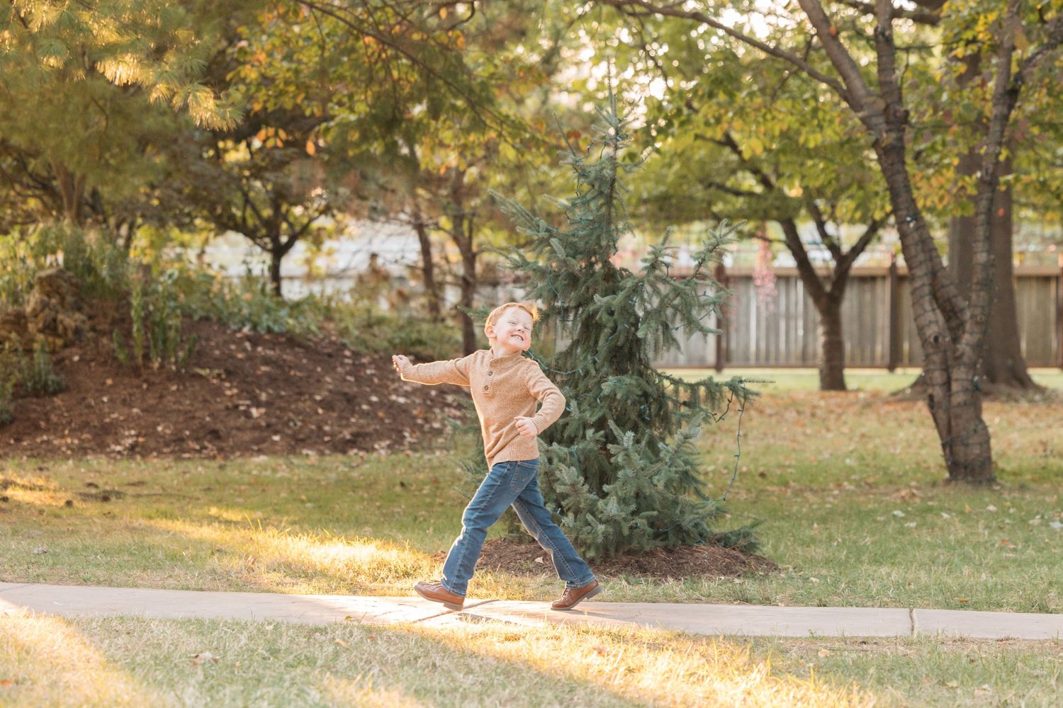 Child jumping along a sunlit sidewalk in fall with trees and a fence in the background.