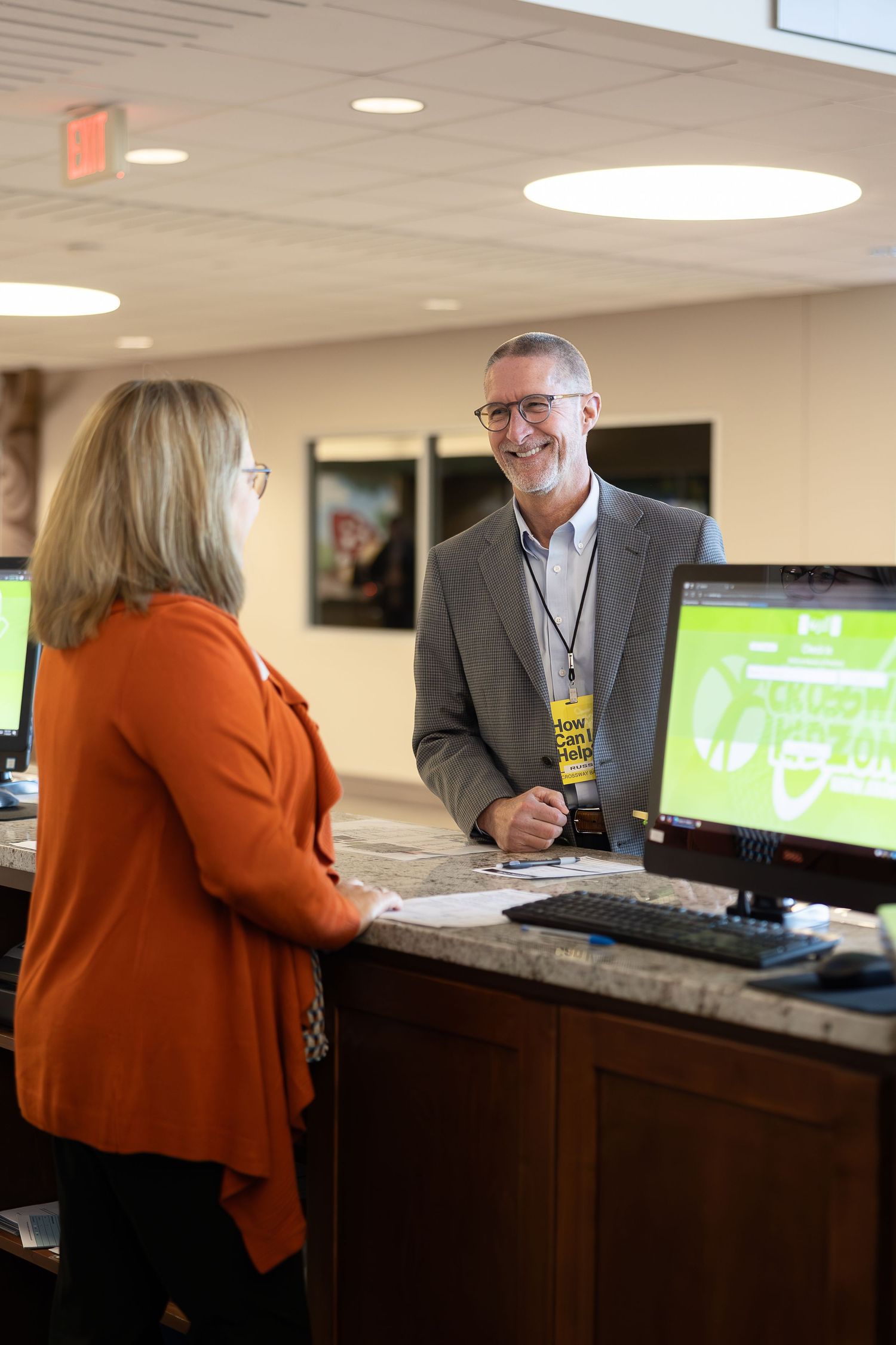 Staff member assists visitor at check-in counter with computer displays showing event information.