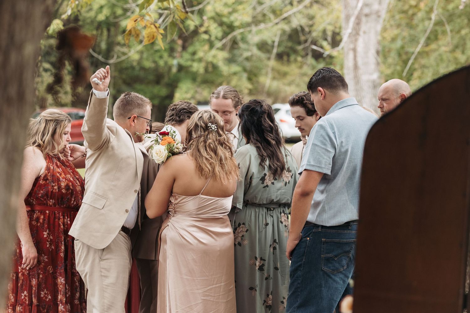 Wedding party gathered outdoors in a wooded setting, with guests in formal attire praying together during a ceremony.