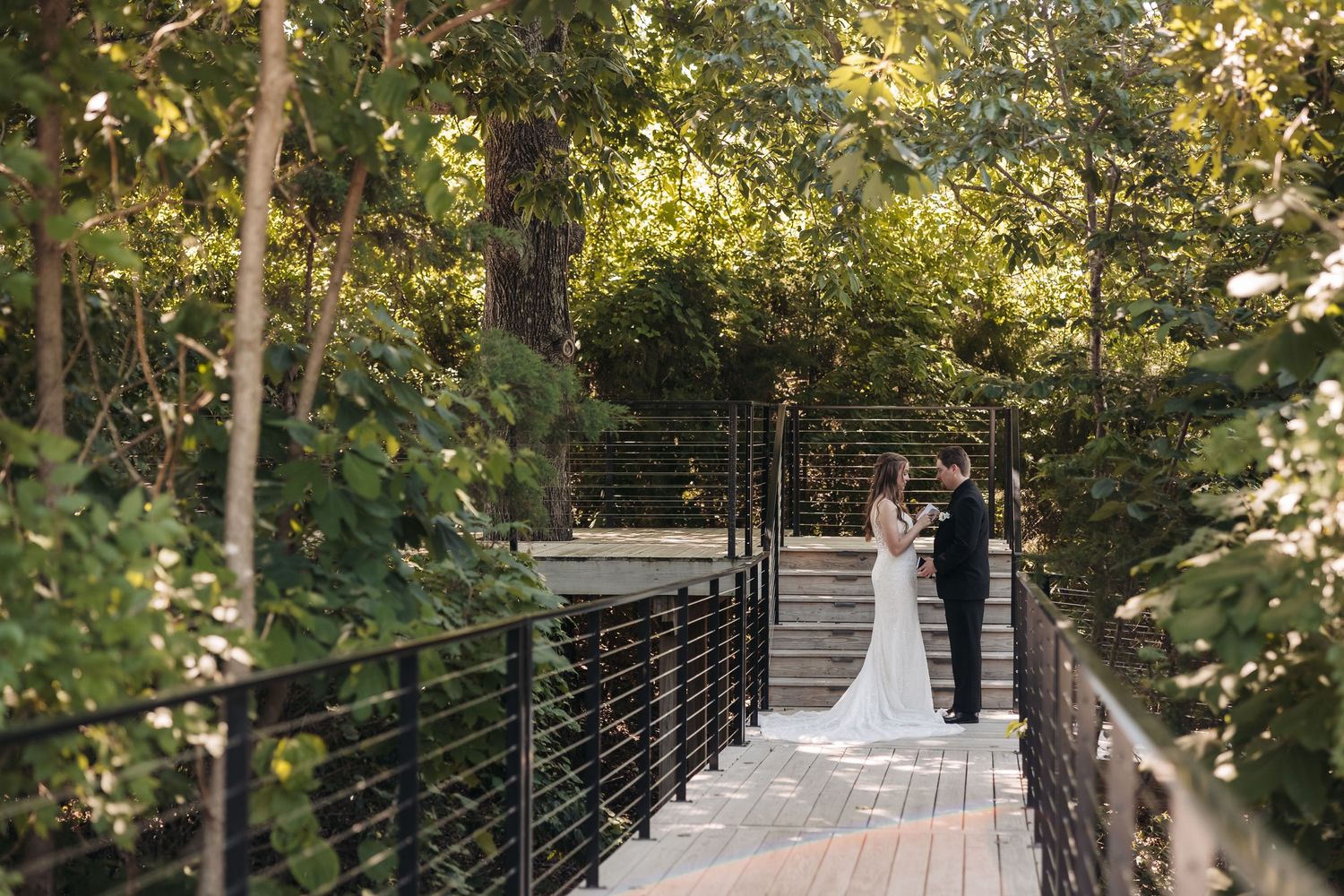 Bride and groom share a romantic moment on a wooden bridge surrounded by lush green trees at an outdoor wedding venue.