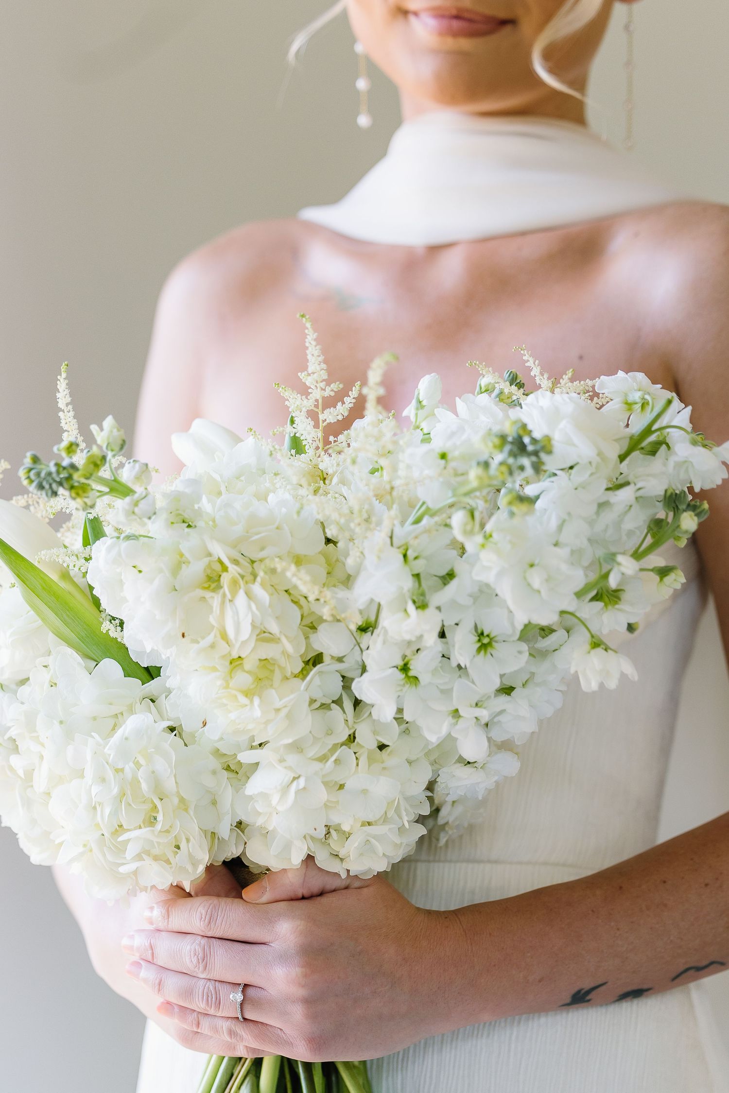 A bride in a white dress holds a stunning bouquet of white delphinium and hydrangea flowers against a light background.