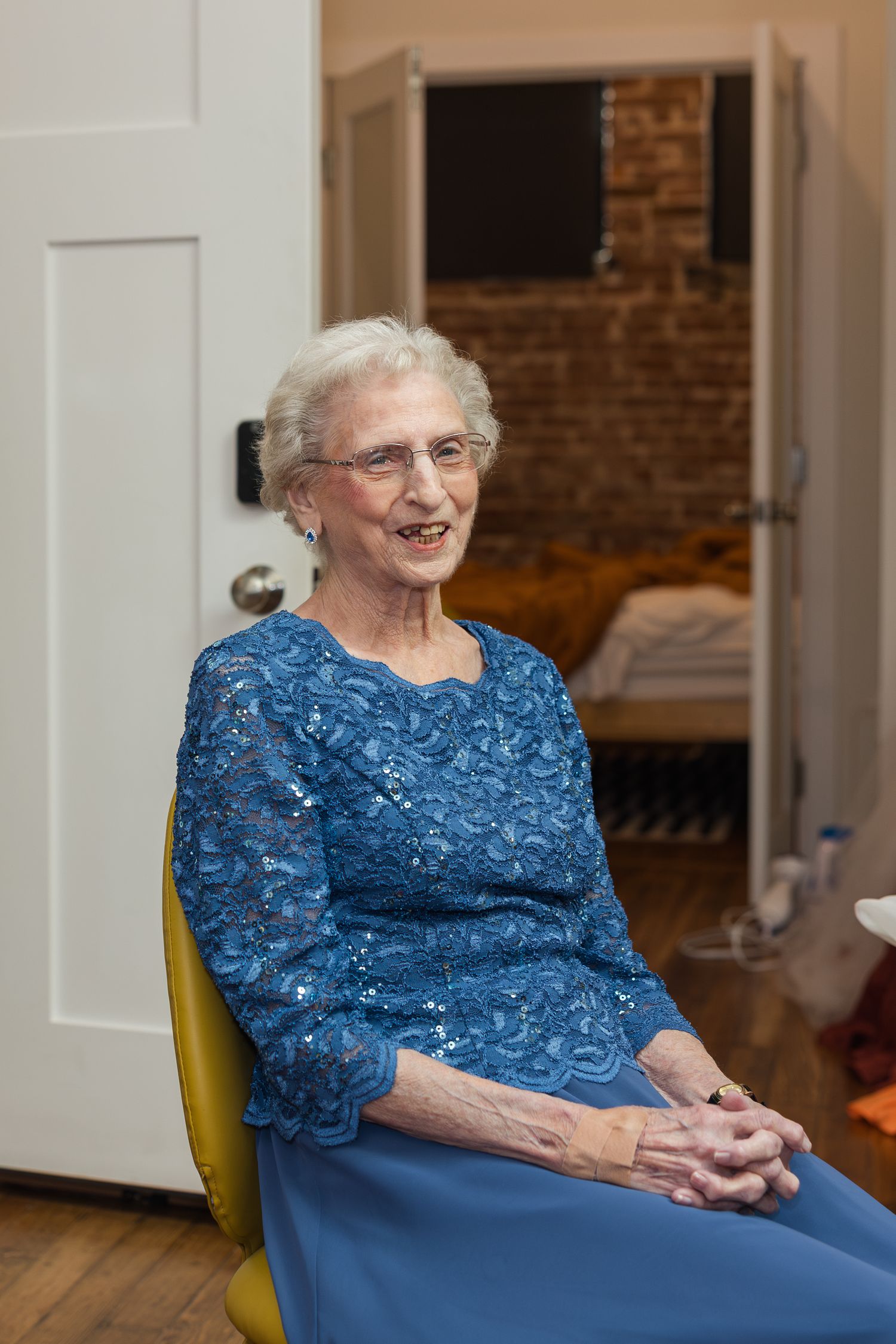 Elderly woman smiling in a blue sequined dress, seated in a yellow chair near an open door.