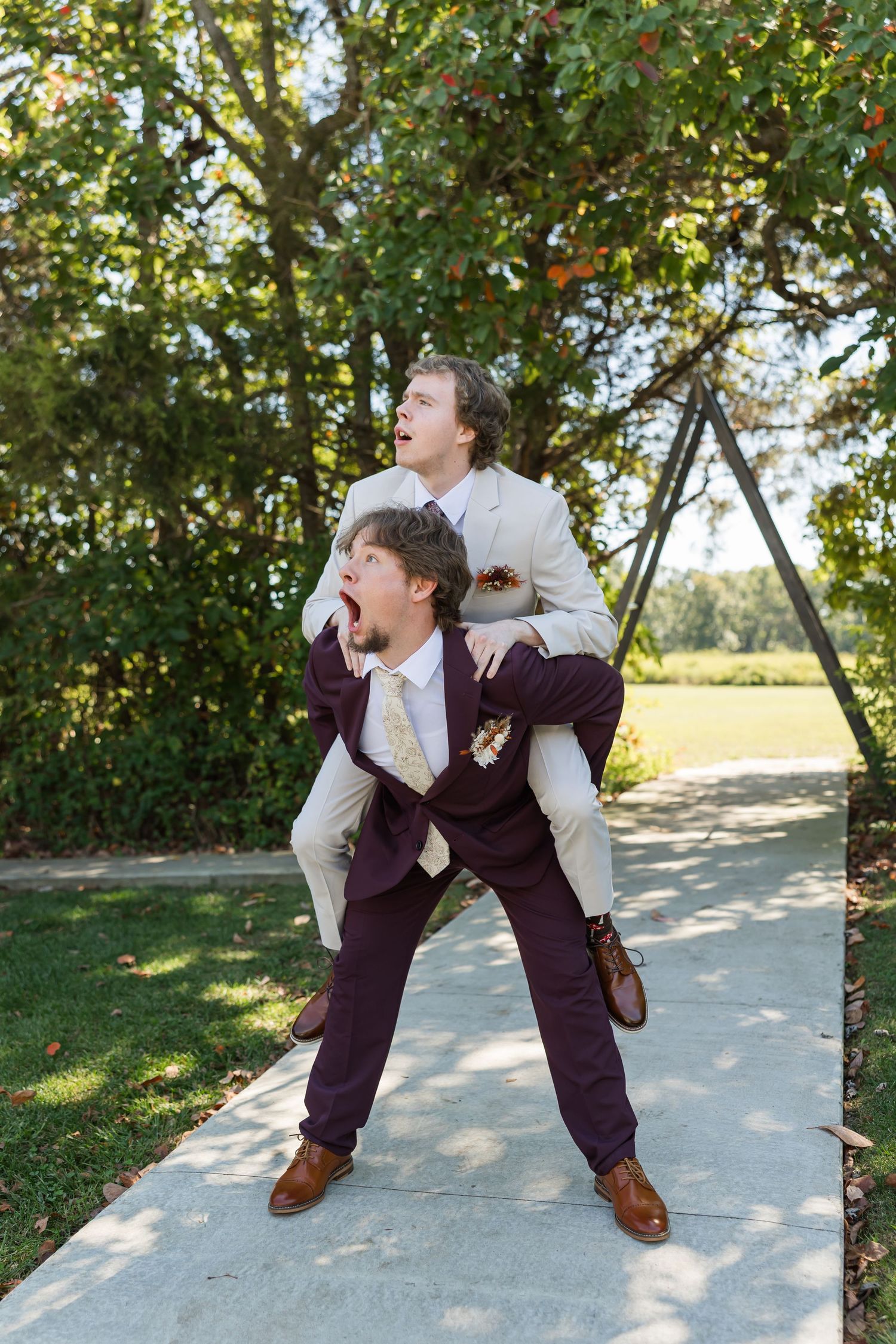 Two men in suits share a playful piggyback ride outdoors, both making exaggerated surprised expressions at a wedding.