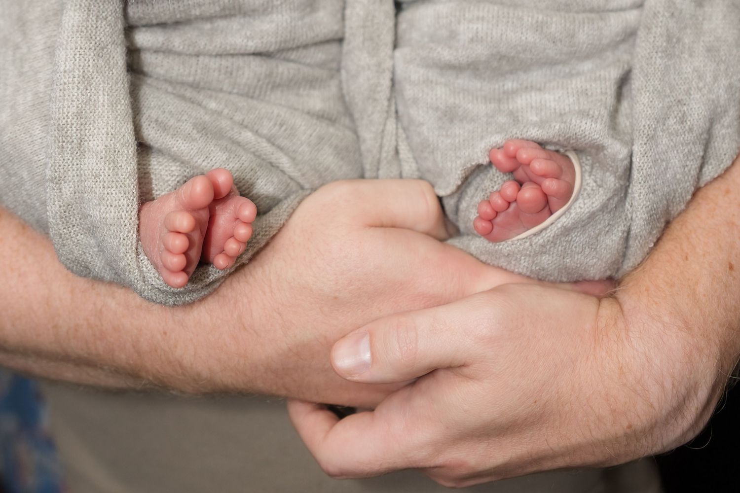 Close-up of a father holding two newborn babies' tiny feet in his hands, wrapped in a gray blanket.