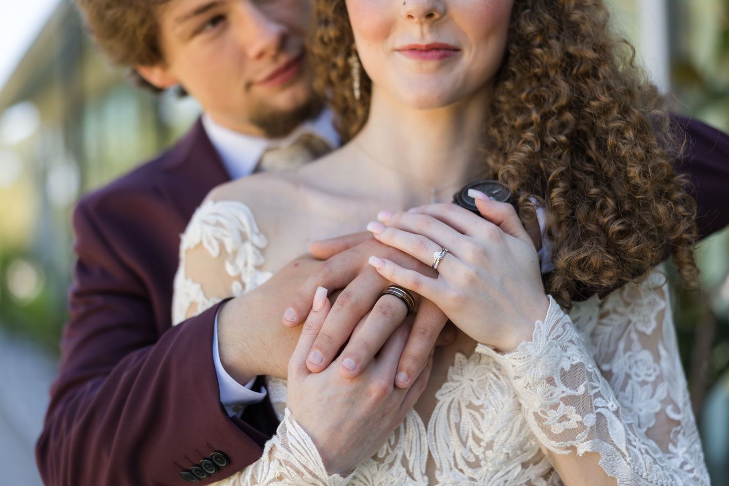 Close up of hands showing off wedding rings with lace dress sleeve and burgundy suit jacket in soft outdoor lighting.