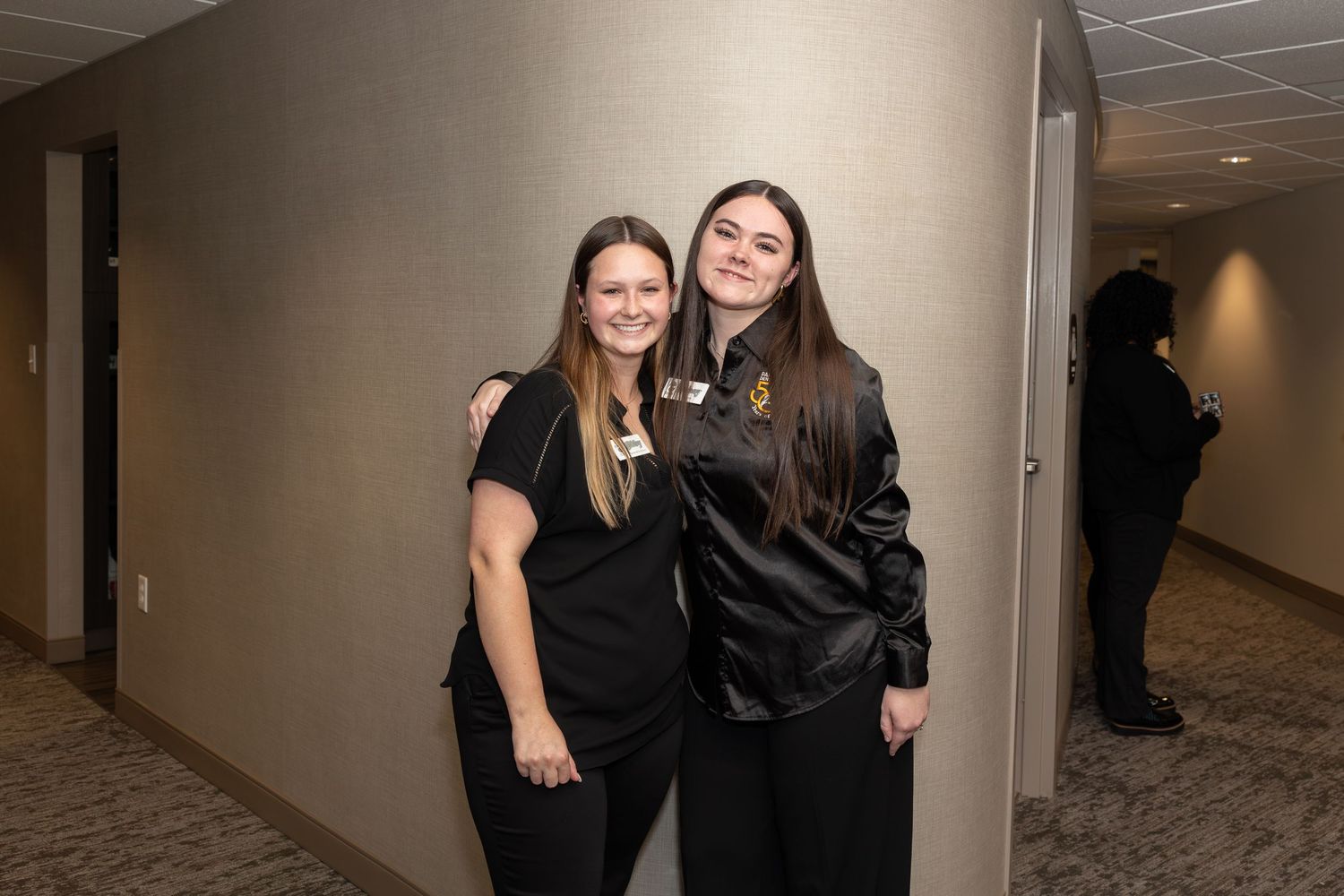 Two colleagues in black attire pose together in an office hallway.