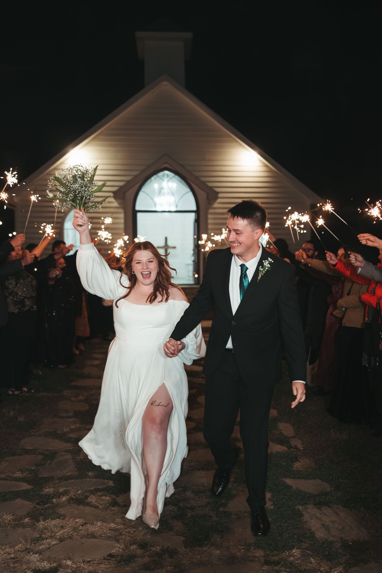 Joyful wedding exit with sparklers outside white chapel at night.