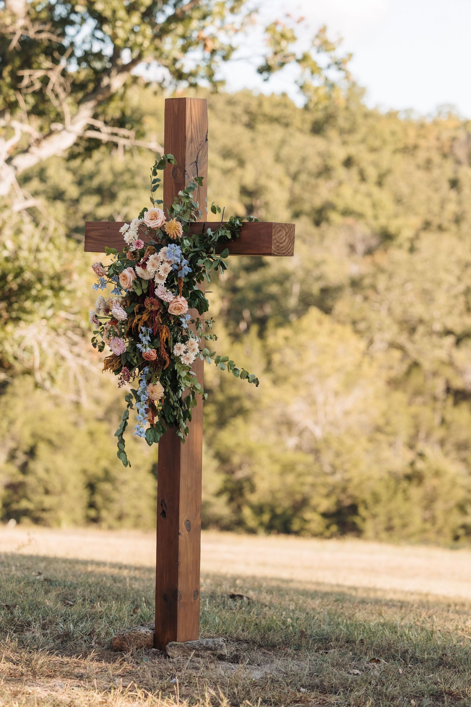 A wooden cross adorned with colorful flowers and greenery stands against a sunlit natural backdrop.