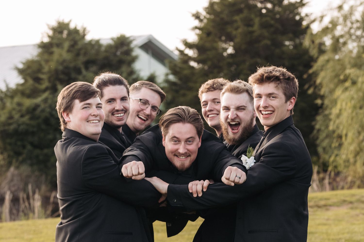 Group of groomsmen in black suits huddle together for fun photo on grassy lawn.