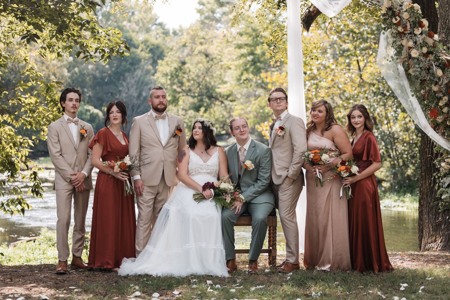 Wedding party in earth-toned attire poses beneath trees during an outdoor autumn ceremony.