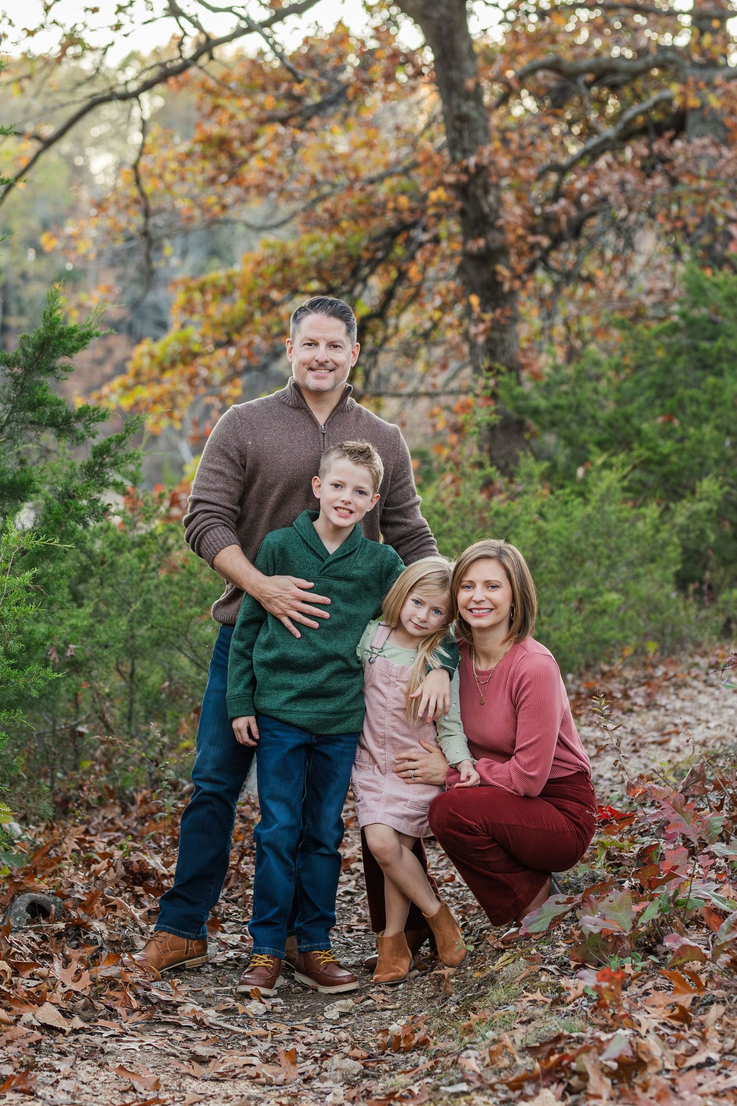 Family in coordinated autumn outfits posing together on a leaf-covered path in a wooded setting.