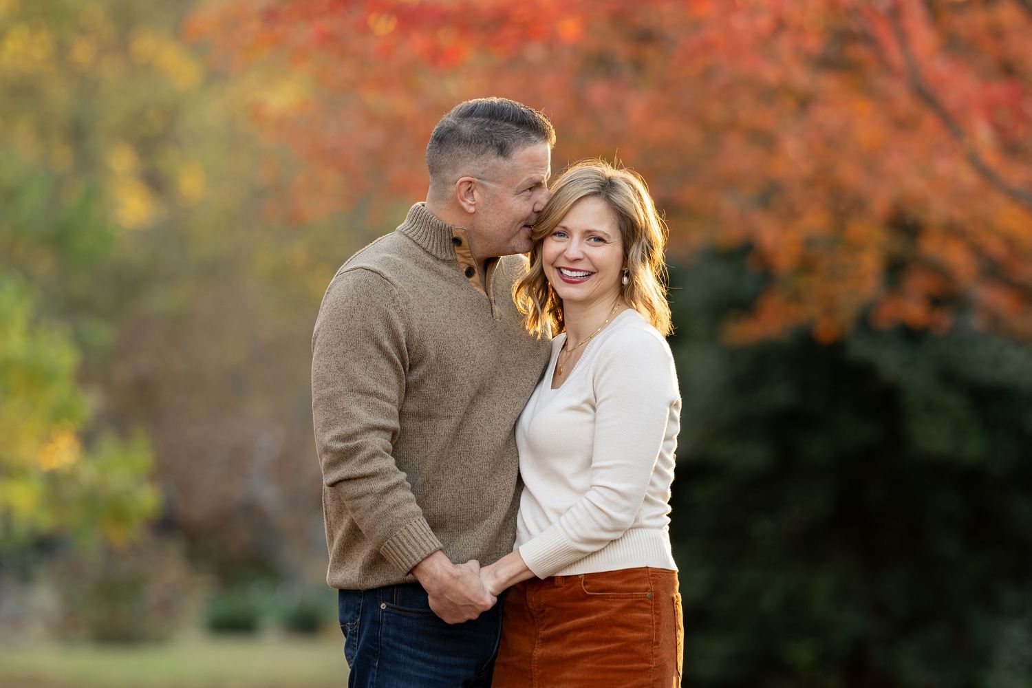 Couple embracing outdoors in autumn, surrounded by vibrant red and orange foliage, wearing cozy sweaters.