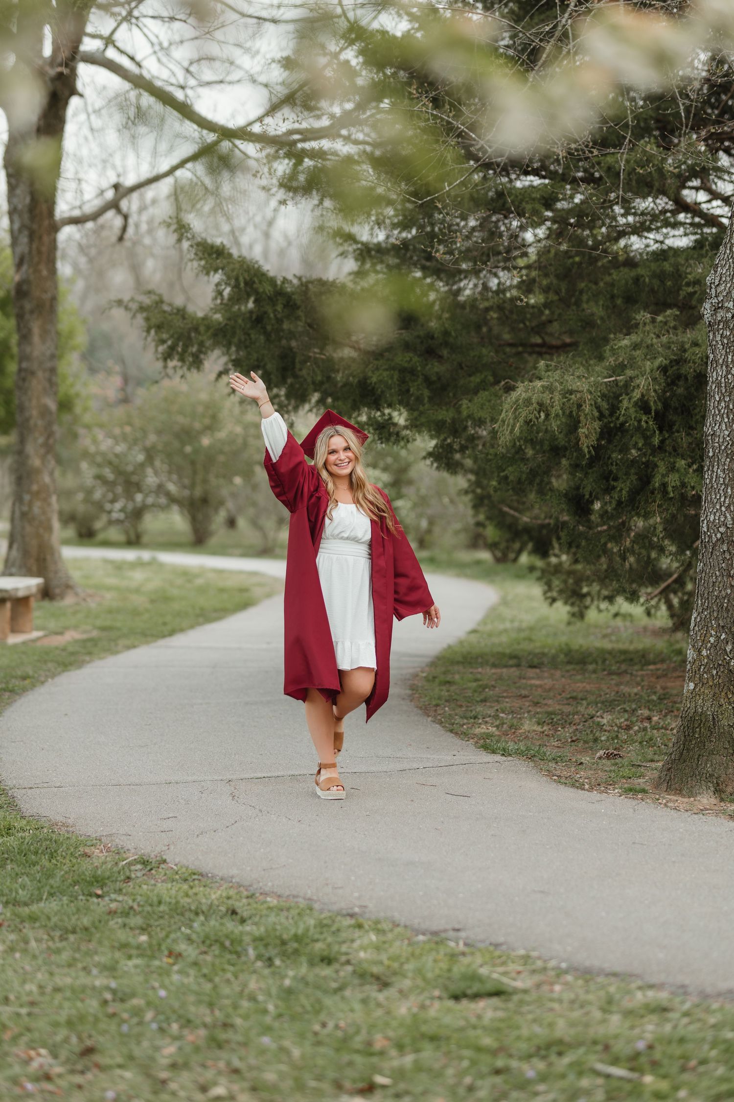 Graduate in red cap and gown celebrates on campus pathway surrounded by trees in springtime.